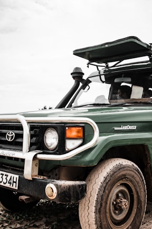A green Toyota Land Cruiser with a visible front bumper and license plate is parked in an outdoor setting. The vehicle has a raised hood, and one person is partially visible inside the driver’s seat.