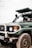 A green Toyota Land Cruiser with a visible front bumper and license plate is parked in an outdoor setting. The vehicle has a raised hood, and one person is partially visible inside the driver’s seat.