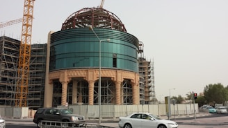 A modern building under construction, featuring a circular glass facade with ornate columns and scaffolding surrounding it. A construction crane is visible in the background, and several vehicles, including a black SUV and a white sedan, are on the road in front of the site. Trees can be seen in the distance under a cloudy sky.