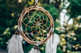 A dreamcatcher with a circular wooden frame is intricately woven with a central web. Blue beads are interspersed throughout the web, and several white feathers hang from the bottom. The background is a blurred natural setting with lush green foliage.