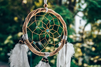 A dreamcatcher with a circular wooden frame is intricately woven with a central web. Blue beads are interspersed throughout the web, and several white feathers hang from the bottom. The background is a blurred natural setting with lush green foliage.