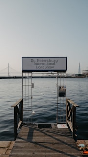 An entrance gate labeled 'St. Petersburg International Boat Show' leads out to calm waters. In the background, a suspension bridge and a modern architectural spire are visible under a clear sky.