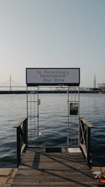 An entrance gate labeled 'St. Petersburg International Boat Show' leads out to calm waters. In the background, a suspension bridge and a modern architectural spire are visible under a clear sky.