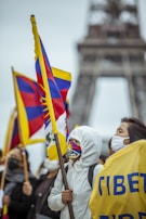 A group of people are gathered for a protest or demonstration, holding flags with Tibetan symbols. Some individuals are wearing masks and protective clothing, possibly indicating a cold or concerned atmosphere. The Eiffel Tower is visible in the background, suggesting the location is Paris.