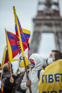 A group of people are gathered for a protest or demonstration, holding flags with Tibetan symbols. Some individuals are wearing masks and protective clothing, possibly indicating a cold or concerned atmosphere. The Eiffel Tower is visible in the background, suggesting the location is Paris.