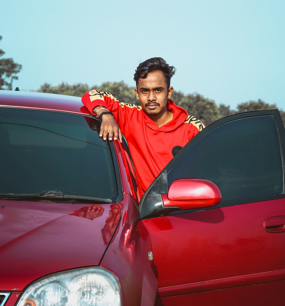 Hombre con sudadera con capucha roja y negra de pie junto a un coche rojo durante el día
