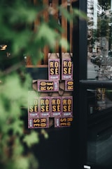 A stack of cardboard containers labeled 'Rose' is displayed near a window. The containers have prominent yellow text on a dark background with some pink decoration. The view is partially obscured by a leafy green plant in the foreground, creating a soft, natural frame around the boxes.