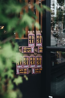 A stack of cardboard containers labeled 'Rose' is displayed near a window. The containers have prominent yellow text on a dark background with some pink decoration. The view is partially obscured by a leafy green plant in the foreground, creating a soft, natural frame around the boxes.
