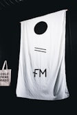 A large white fabric backdrop with minimalist black symbols hangs against a dark background. In the lower left corner, a beige tote bag with the text 'COLORADO' in black capital letters, split into three lines with a symbol replacing the letter 'I'.