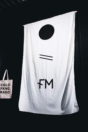 A large white fabric backdrop with minimalist black symbols hangs against a dark background. In the lower left corner, a beige tote bag with the text 'COLORADO' in black capital letters, split into three lines with a symbol replacing the letter 'I'.