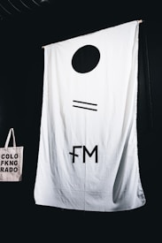 A large white fabric backdrop with minimalist black symbols hangs against a dark background. In the lower left corner, a beige tote bag with the text 'COLORADO' in black capital letters, split into three lines with a symbol replacing the letter 'I'.