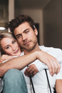 A close-up of two people sitting closely together, exuding a sense of warmth and intimacy. One person has short brown hair and a beard, while the other person has long blonde hair. Both are wearing casual clothing and appear relaxed.