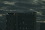 A tall hotel building with the name 'Grand Hyatt' lit up on its top, set against a cloudy, dark sky. The building's windows have faint lights visible, suggesting it is evening or night.