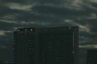 A tall hotel building with the name 'Grand Hyatt' lit up on its top, set against a cloudy, dark sky. The building's windows have faint lights visible, suggesting it is evening or night.
