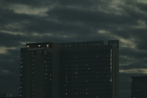 A tall hotel building with the name 'Grand Hyatt' lit up on its top, set against a cloudy, dark sky. The building's windows have faint lights visible, suggesting it is evening or night.