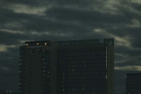 A tall hotel building with the name 'Grand Hyatt' lit up on its top, set against a cloudy, dark sky. The building's windows have faint lights visible, suggesting it is evening or night.
