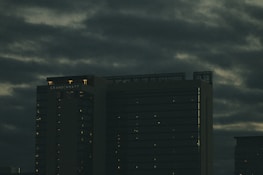 A tall hotel building with the name 'Grand Hyatt' lit up on its top, set against a cloudy, dark sky. The building's windows have faint lights visible, suggesting it is evening or night.