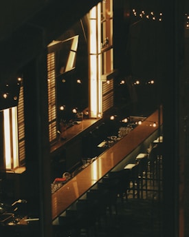 A dimly lit bar area featuring a long polished counter with several chairs neatly arranged. Warm, ambient lighting emanates from vertical fixtures and string lights, creating a cozy and intimate atmosphere. Glasses and bottles are visible on the counter, suggesting a ready-to-serve setup.