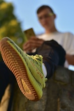 A person is sitting with one foot forward wearing a pair of yellow sneakers with distinctive red lettering on the side. The background is blurred, focusing on the shoe's design and texture.