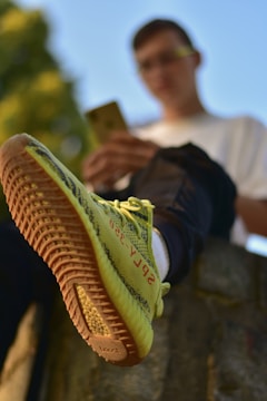 A person is sitting with one foot forward wearing a pair of yellow sneakers with distinctive red lettering on the side. The background is blurred, focusing on the shoe's design and texture.