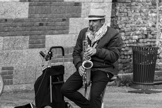 A person wearing a patterned hat and scarf plays a saxophone while seated on an outdoor stool. A rolling cart with various items rests nearby, alongside a woven trash can. The background features a textured brick wall with alternating stone patterns.