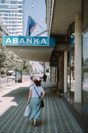 A person wearing a white top and green skirt is walking down a patterned sidewalk in an urban area. The area features a bank sign with the name 'ABANKA', and there are modern buildings and trees lining the street. The person is carrying a white bag and has a crossbody strap over their shoulder.