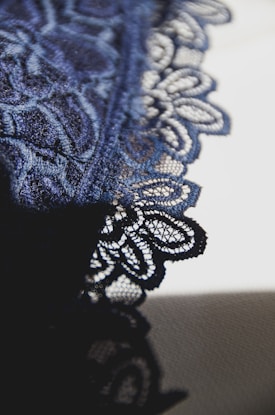 Intricate lace fabric with detailed floral patterns in a deep blue shade is displayed, revealing the fine texture and craftsmanship. The fabric is set against a contrasting light background, emphasizing the delicate design.