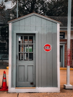 A small wooden structure with a gray door and siding, featuring a 'Gulf' logo sign on the front. The structure is located outdoors, next to a traffic cone on the left and a 'No Smoking' sign on the right. The background includes parts of a brick building and some trees.