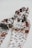 A patterned textile scarf in shades of cream, brown, and black, intricately twisted together, set against a plain white background, creating a visually appealing design.