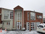 A two-story building with large windows is surrounded by snow. The structure is constructed with a mix of light-colored and wooden panels. A sign displaying 'HAKA LODGE Backpackers' is visible on the right side. Snow is actively falling, and a white vehicle is parked in the front with traffic cones placed nearby.