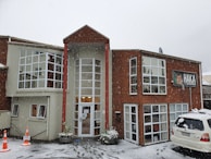 A two-story building with large windows is surrounded by snow. The structure is constructed with a mix of light-colored and wooden panels. A sign displaying 'HAKA LODGE Backpackers' is visible on the right side. Snow is actively falling, and a white vehicle is parked in the front with traffic cones placed nearby.