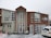 A two-story building with large windows is surrounded by snow. The structure is constructed with a mix of light-colored and wooden panels. A sign displaying 'HAKA LODGE Backpackers' is visible on the right side. Snow is actively falling, and a white vehicle is parked in the front with traffic cones placed nearby.