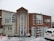 A two-story building with large windows is surrounded by snow. The structure is constructed with a mix of light-colored and wooden panels. A sign displaying 'HAKA LODGE Backpackers' is visible on the right side. Snow is actively falling, and a white vehicle is parked in the front with traffic cones placed nearby.