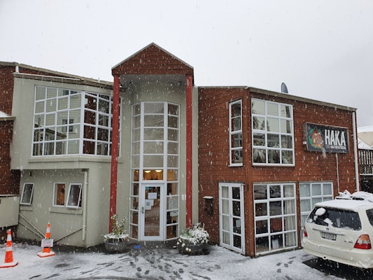 A two-story building with large windows is surrounded by snow. The structure is constructed with a mix of light-colored and wooden panels. A sign displaying 'HAKA LODGE Backpackers' is visible on the right side. Snow is actively falling, and a white vehicle is parked in the front with traffic cones placed nearby.
