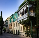 A row of charming, historic-style buildings with green roofs and ornate white balconies is adorned with lush ivy. The architectural design features arched windows and detailed woodwork. The street is paved with cobblestones and lined with tall, slender trees.