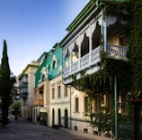 A row of charming, historic-style buildings with green roofs and ornate white balconies is adorned with lush ivy. The architectural design features arched windows and detailed woodwork. The street is paved with cobblestones and lined with tall, slender trees.