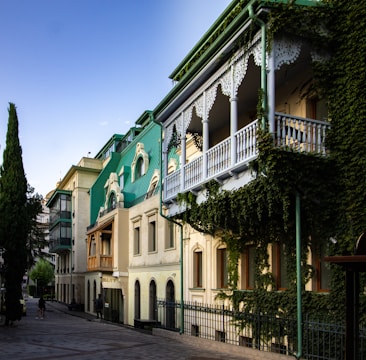 A row of charming, historic-style buildings with green roofs and ornate white balconies is adorned with lush ivy. The architectural design features arched windows and detailed woodwork. The street is paved with cobblestones and lined with tall, slender trees.