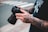 A person with heavily tattooed arms holds a professional camera with both hands. The tattoos are intricate and cover most of the visible skin. The person is wearing a black shirt with a graphic design. The scene appears to be outdoors, with a blurred background indicating a street crossing.
