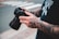 A person with heavily tattooed arms holds a professional camera with both hands. The tattoos are intricate and cover most of the visible skin. The person is wearing a black shirt with a graphic design. The scene appears to be outdoors, with a blurred background indicating a street crossing.