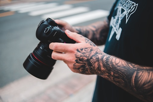 A person with heavily tattooed arms holds a professional camera with both hands. The tattoos are intricate and cover most of the visible skin. The person is wearing a black shirt with a graphic design. The scene appears to be outdoors, with a blurred background indicating a street crossing.