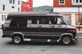 A classic brown van is driving on a street, passing by colorful buildings, including a red and a white one. The van has a retro design with tinted windows and chrome accents. The background features a glimpse of a hillside under a cloudy sky.