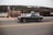 A black classic car is driving along a road in an urban area. The scene includes a fenced area on the left with a large hill in the background. There are some industrial elements visible, such as a truck and a fire hydrant.
