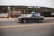 A black classic car is driving along a road in an urban area. The scene includes a fenced area on the left with a large hill in the background. There are some industrial elements visible, such as a truck and a fire hydrant.