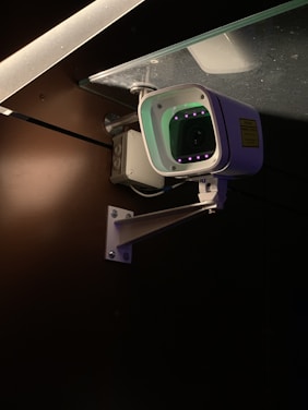 A security camera is mounted on a metal bracket attached to a wall. The camera is positioned under a glass surface with visible screws and brackets. The environment is dimly lit, highlighting the camera's body and the glowing infrared lights around the lens.