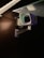 A security camera is mounted on a metal bracket attached to a wall. The camera is positioned under a glass surface with visible screws and brackets. The environment is dimly lit, highlighting the camera's body and the glowing infrared lights around the lens.