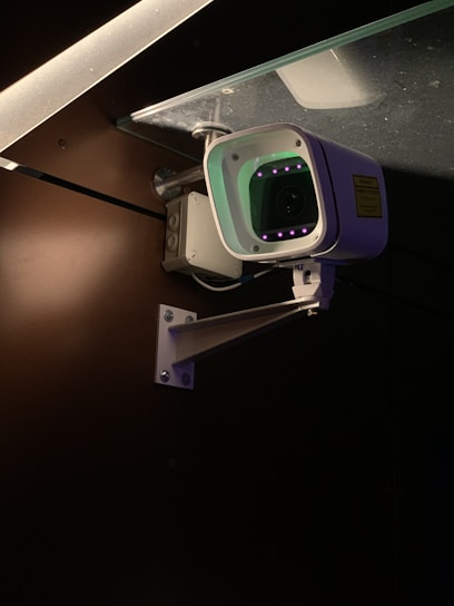 A security camera is mounted on a metal bracket attached to a wall. The camera is positioned under a glass surface with visible screws and brackets. The environment is dimly lit, highlighting the camera's body and the glowing infrared lights around the lens.