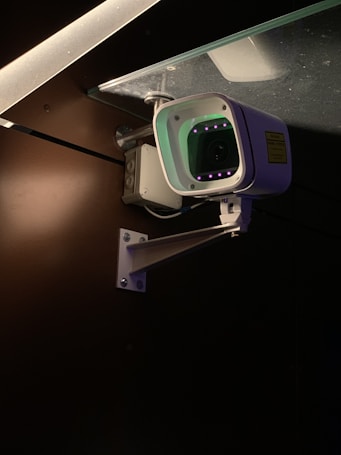 A security camera is mounted on a metal bracket attached to a wall. The camera is positioned under a glass surface with visible screws and brackets. The environment is dimly lit, highlighting the camera's body and the glowing infrared lights around the lens.