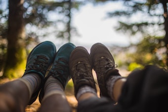 A pair of feet wearing hiking shoes are stretched out against a blurred background of forest greenery and sunlight. The focus is on the shoes, one pair is teal and the other is brown, both atop patterned socks.