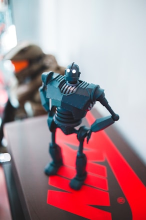 A small robot figure stands on a surface with a bright red logo on it, showing mechanical joints, a cylindrical head with glowing eyes, and detailed body features.