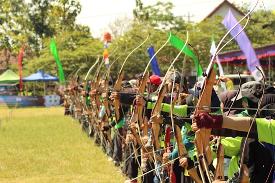A group of archers stands in a line, each holding a bow and aiming at a target. They are outdoors, and the setting includes multiple colorful flags mounted in the background. The archers appear to be participating in a coordinated event, characterized by the uniformity of their actions and the diversity of their clothing, which adds vibrancy to the scene.